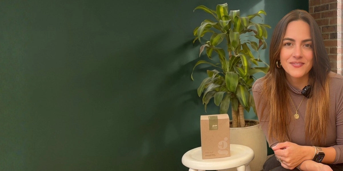 Sara sonriendo frente a un fondo verde oscuro con una planta de interior, acompañada de una caja de empaque ecológico Gimo apoyada sobre un banco blanco.